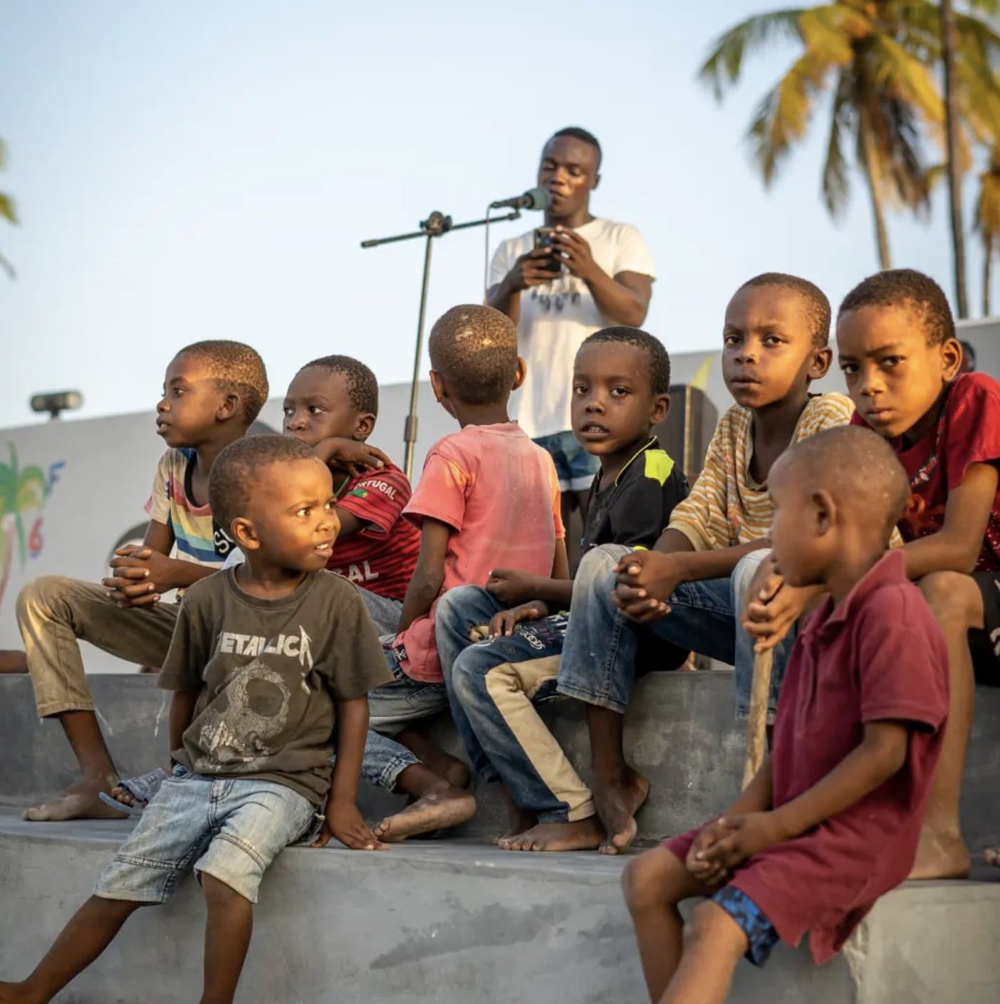 Children from the Move Zanzibar community sitting together, listening to a young performer with a microphone, palm trees behind them Children from the Move Zanzibar community sitting together, listening to a young performer with a microphone, palm trees behind them
